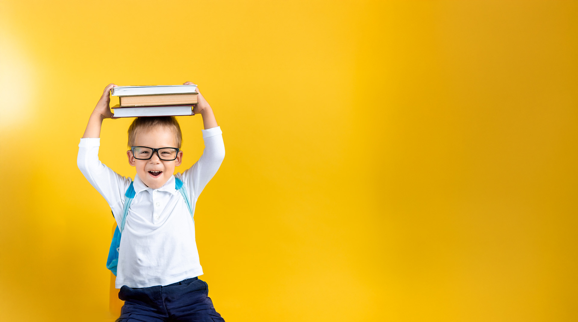 Photo d'un enfant avec des lunettes, une chemise et un sac à dos portant des livres au dessus de sa tête